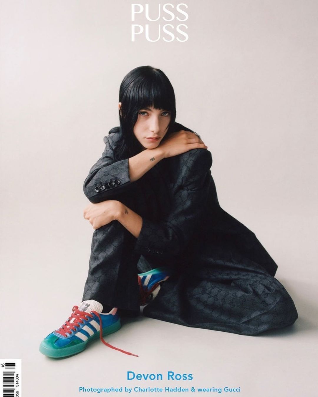 Portrait of Devon Ross wearing a gray suit and sneakers, sitting in front of a white backdrop for Puss Puss magazine.
