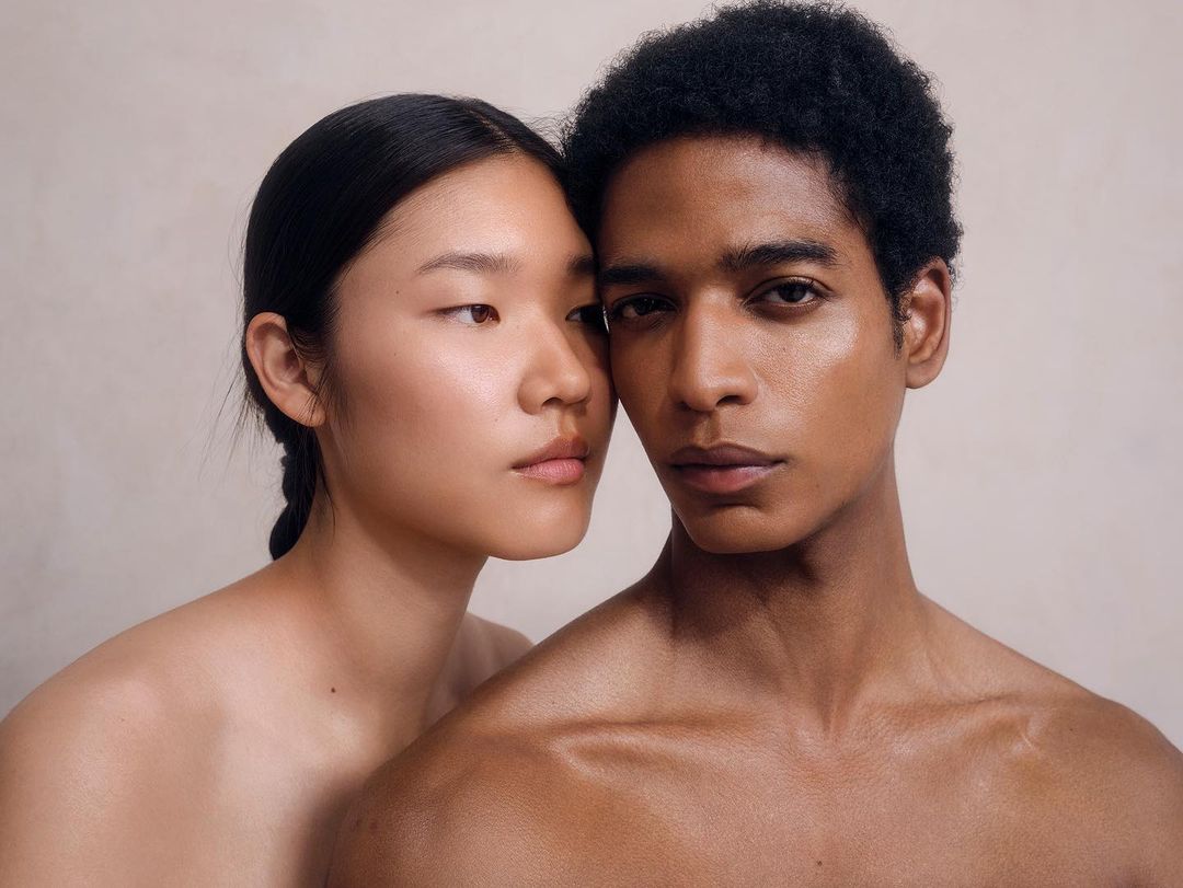 Portrait of a stylish couple with dark hair and light skin posing in front of a plain neutral wall in a beauty or fashion style shot.