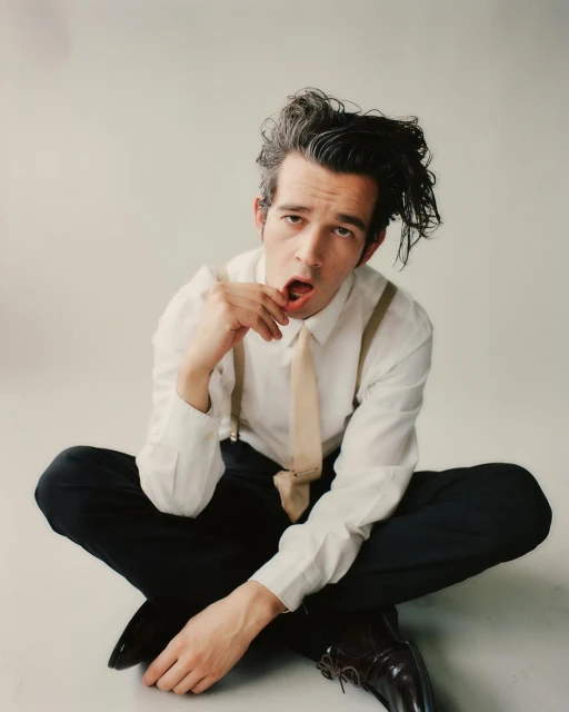 Adult male seated with open mouth and hand to face. He is wearing a light shirt with tan tie and suspenders against a neutral backdrop.