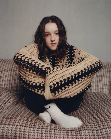 Young person sitting cross-legged on a patterned sofa wearing an oversized cream and black knit sweater
