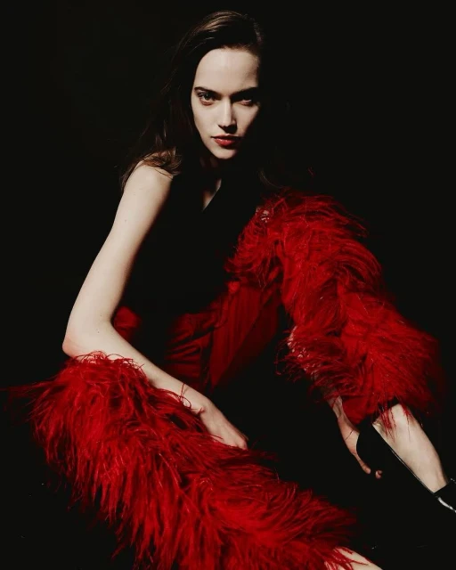 Brunette woman in black top and red feathered pants sits in dark studio, gazing intensely at the viewer in a fashion pose.