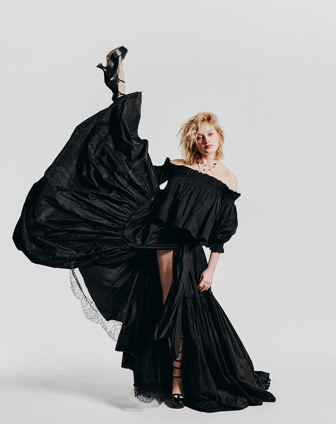 Fashion shot of a blonde woman wearing an off-the-shoulder black gown, one leg raised with black heel, set against a white backdrop.