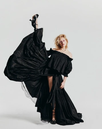Fashion shot of a blonde woman wearing an off-the-shoulder black gown, one leg raised with black heel, set against a white backdrop.