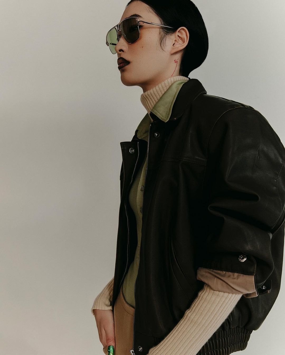 Portrait of an adult woman with dark lipstick, wearing a black beret, aviator sunglasses and a black leather jacket indoors.