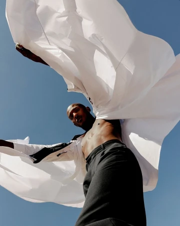 Low angle shot of a man in dark trousers holding a billowing white shirt against a clear blue sky by Charlotte Navio