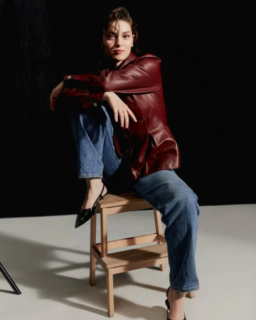Brunette woman in a burgundy leather jacket and jeans poses seated on a wooden step stool in a photo studio with a black backdrop.