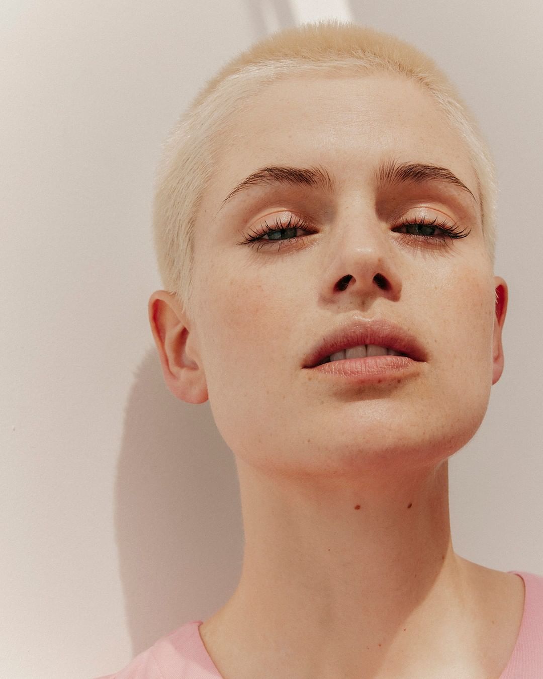 Close-up portrait of a woman with short blonde buzzcut hair wearing a pink top against a neutral background by Charlotte Navio