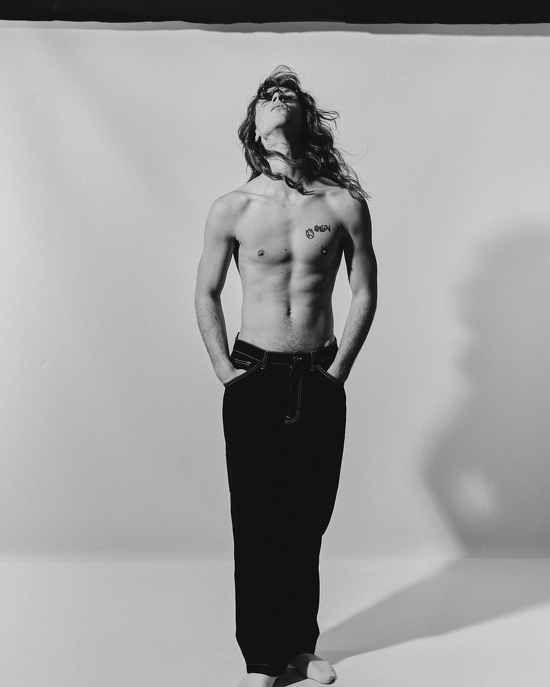 Shirtless man with long hair and dark pants standing with hands in pockets against a plain studio background.