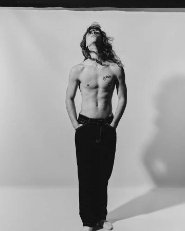 Shirtless man with long hair and dark pants standing with hands in pockets against a plain studio background.