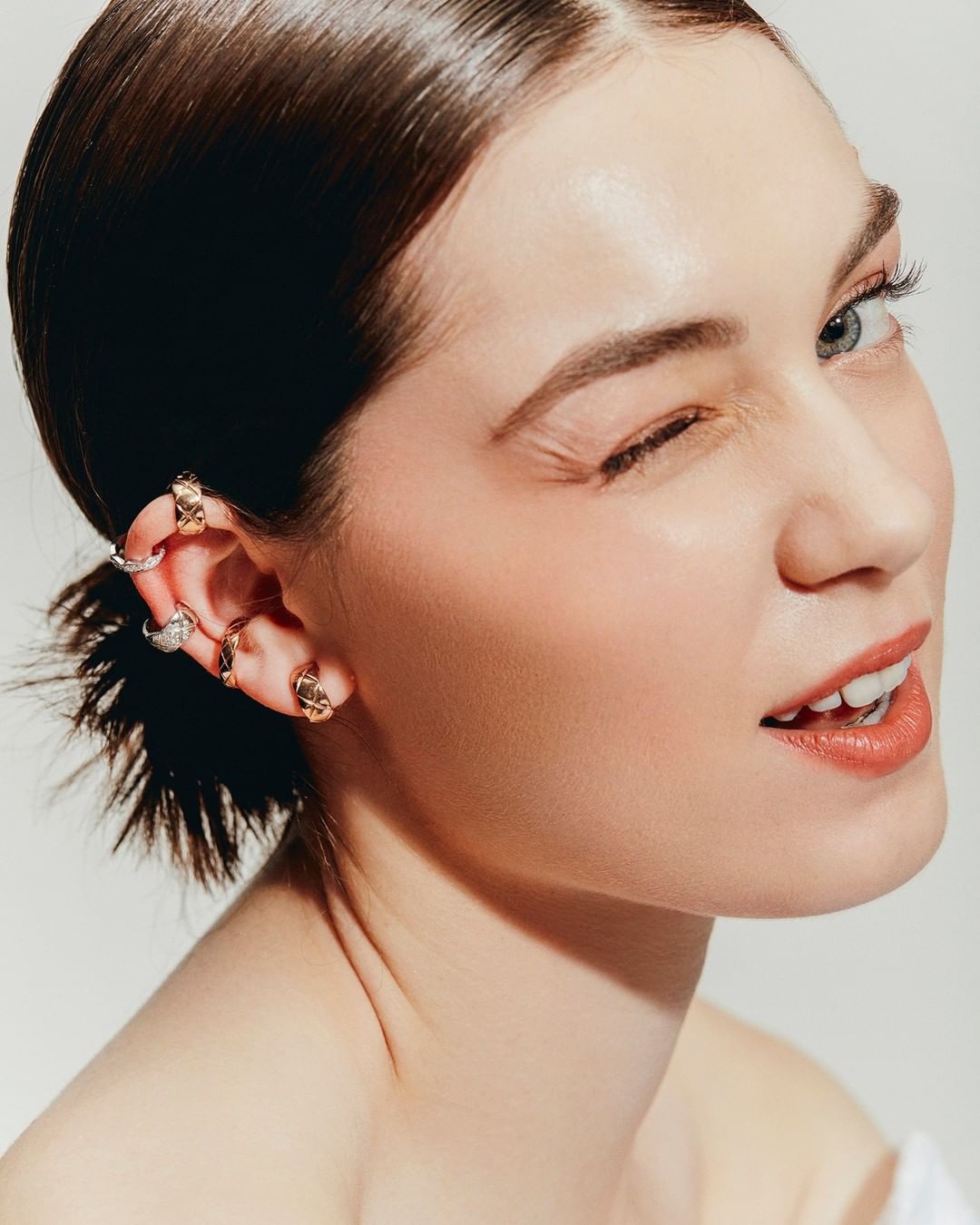 Close-up of a young woman with slicked-back hair winking at the camera, wearing multiple ear cuff rings on one ear.