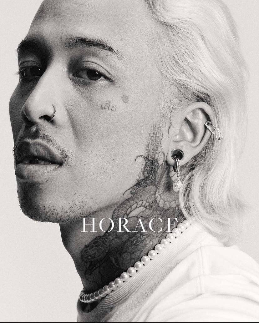 Close-up black and white portrait of an adult male with tattoos, facial piercings, light hair, and pearl necklace against a light background.