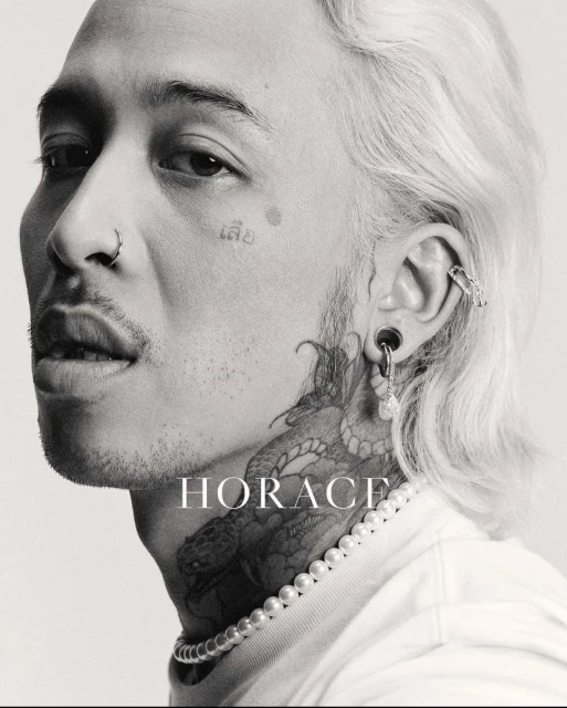 Close-up black and white portrait of an adult male with tattoos, facial piercings, light hair, and pearl necklace against a light background.