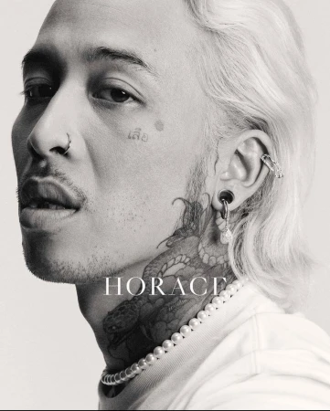 Close-up black and white portrait of an adult male with tattoos, facial piercings, light hair, and pearl necklace against a light background.