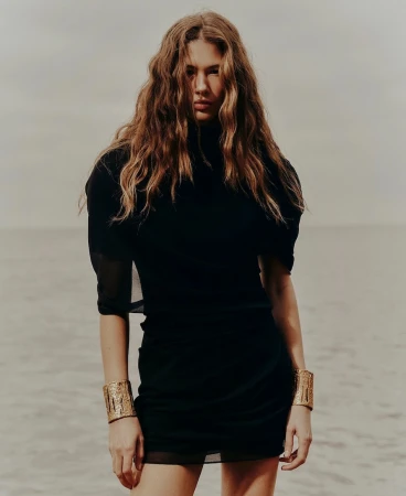 Woman with long wavy brown hair wearing a black dress and gold cuffs against an overcast sky and sandy beach