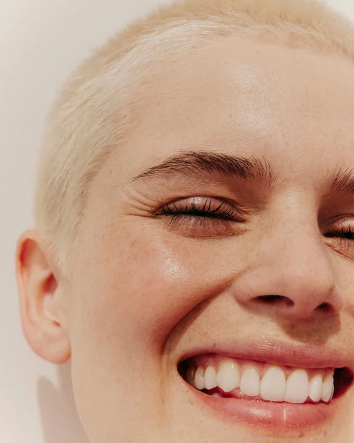 Close-up portrait of a smiling young woman with short blonde hair and closed eyes against a neutral background, showcasing a cheerful expression.