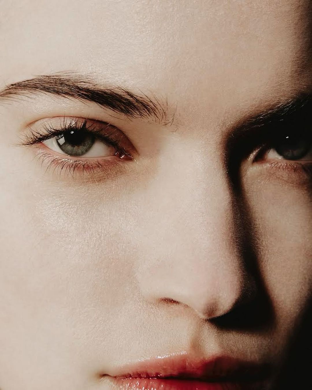 Close-up portrait of a fair-skinned person with prominent eyebrows, green eyes, and glossy lips. Light to medium lighting emphasizes skin texture.