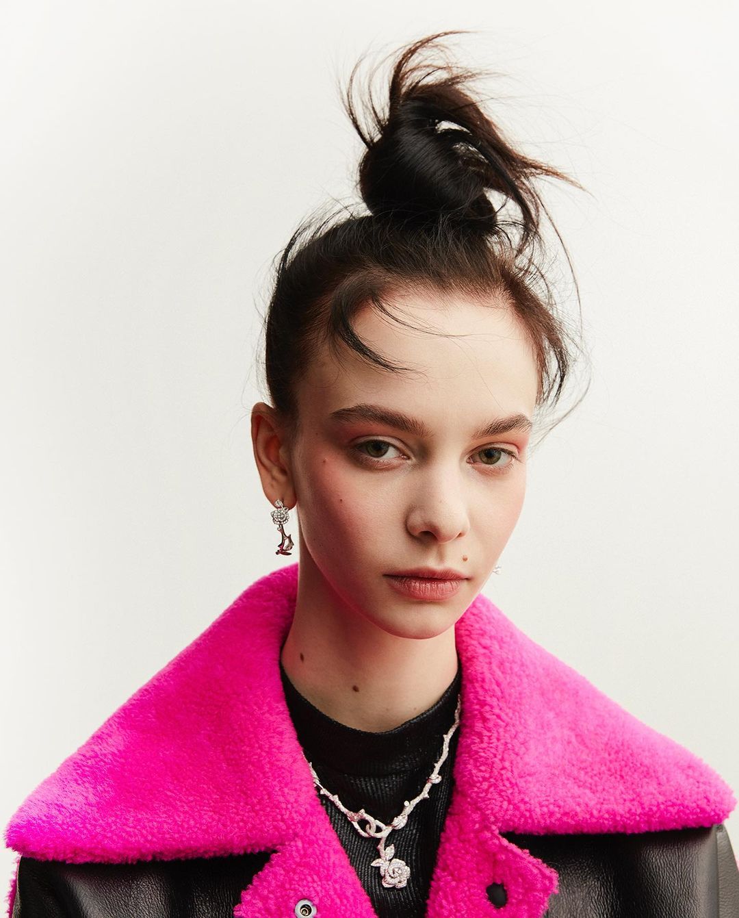 Portrait of a young woman in a hot pink faux fur collar jacket with silver rose jewelry, set against a bright white background.