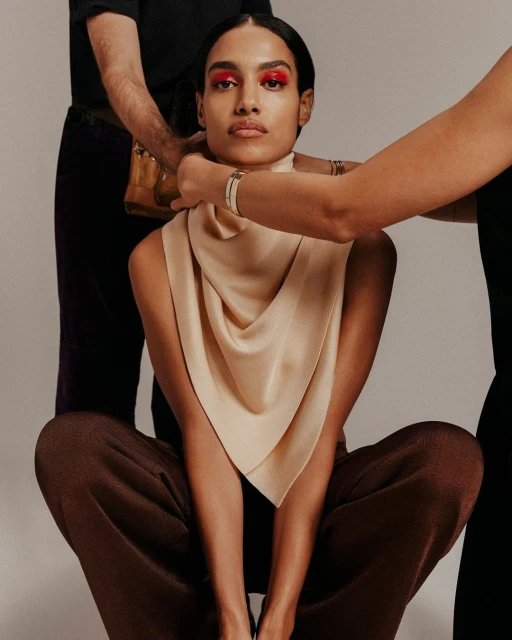 Headshot of a model in an indoor photo studio wearing a beige draped garment and dark brown pants, as two people adjust the fabric.
