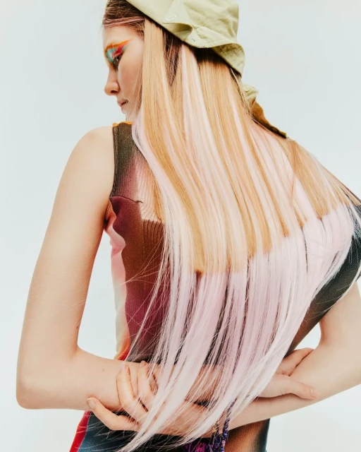 An adult female model with pink-blonde highlighted hair, wearing a beige bucket hat and colorful top, posing in a brightly lit studio.