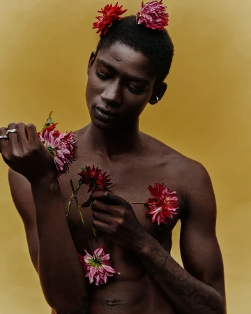 Portrait of a Black man with short hair and red flowers tucked in his hair and chest, posed against a warm mustard yellow backdrop