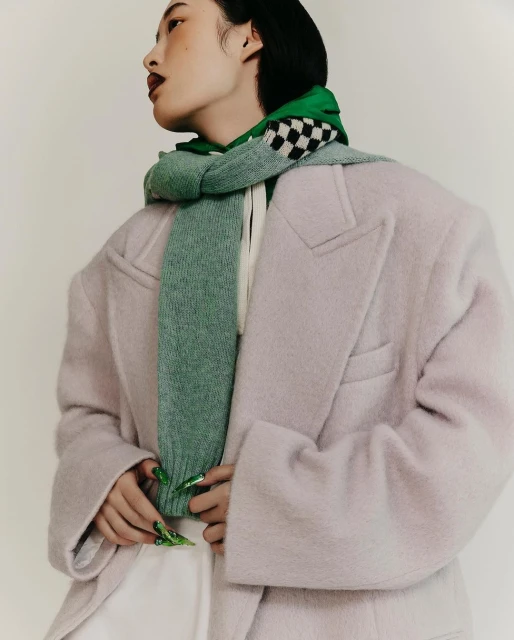 Stylish woman in a pink coat and green scarf poses indoors. Close-up of a person wearing fashionable outerwear in a neutral studio setting.