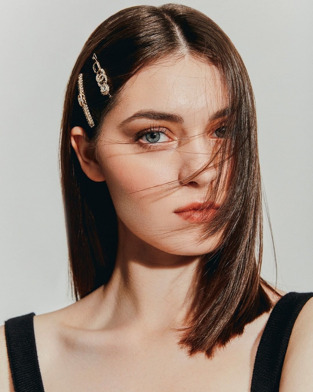 Close-up portrait of a young woman with brunette hair styled with jeweled hair clips, against a neutral studio backdrop in soft lighting.