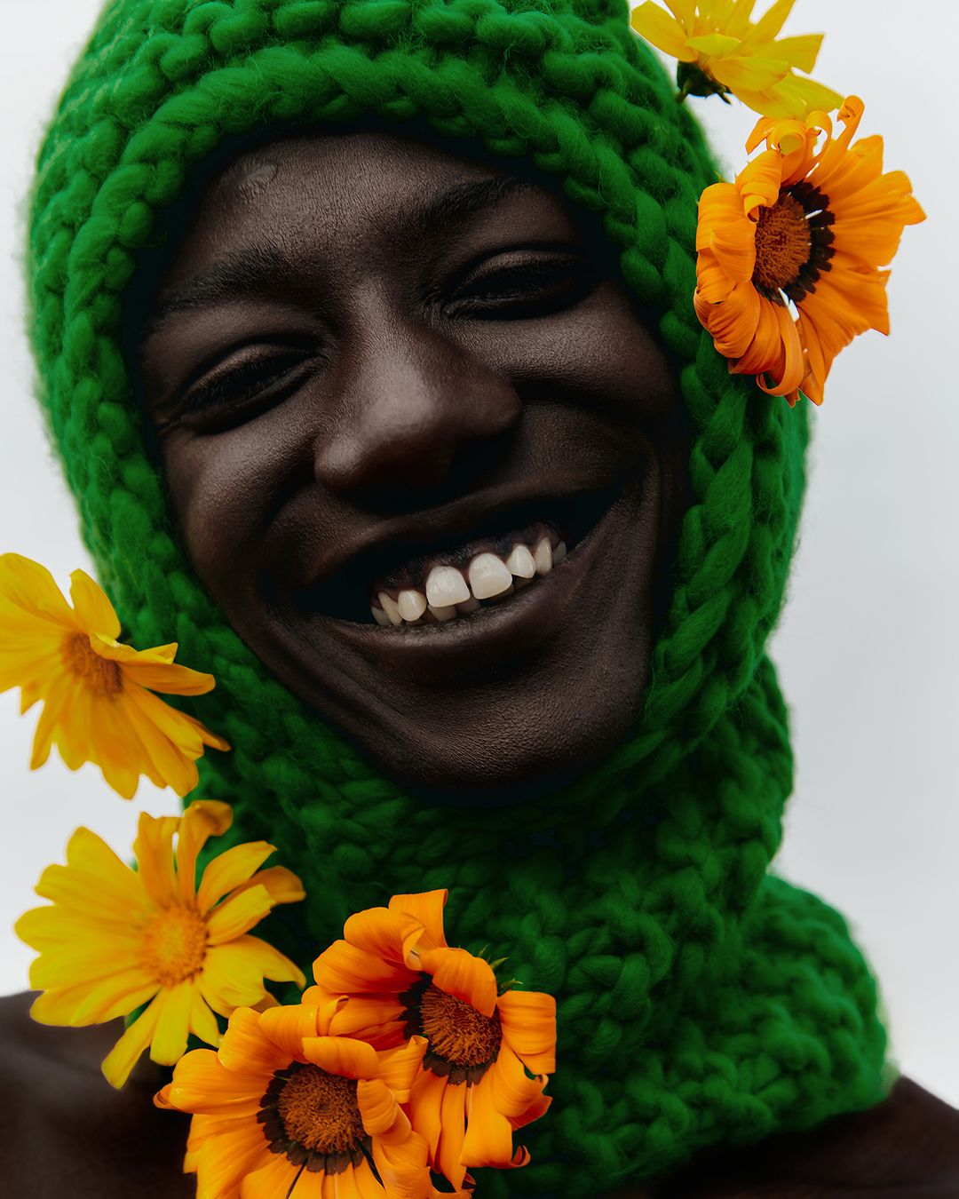 Close-up portrait of a smiling African American adult, wearing a green knit balaclava adorned with yellow and orange flowers. Beauty shot.
