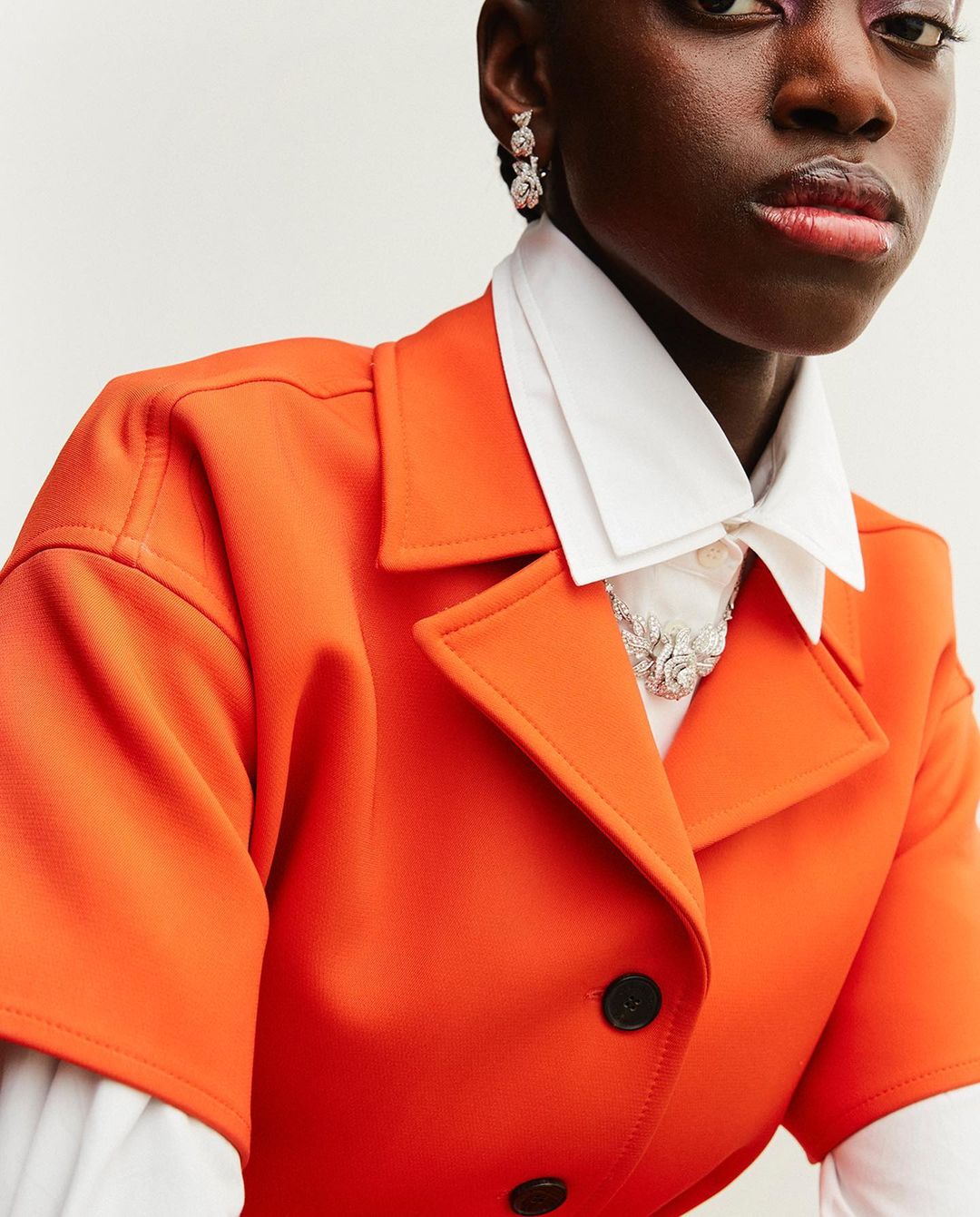 Close-up of a stylish African-American woman in an orange jacket, white collared shirt, and jewel earrings against a white backdrop.