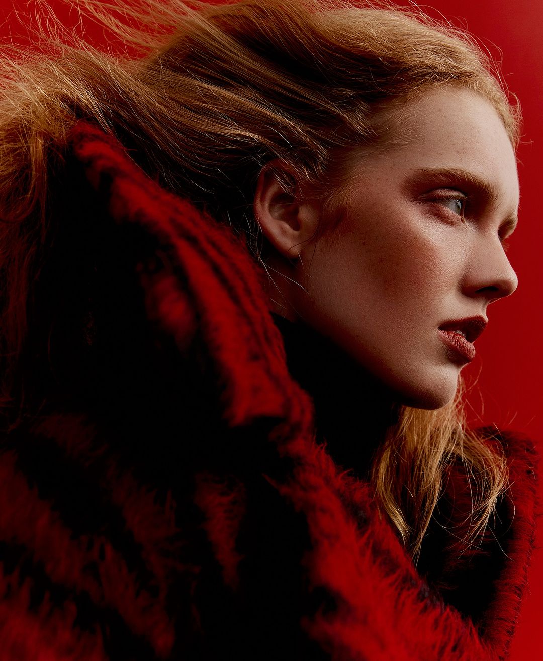 Profile portrait of a fair-skinned adult woman with light hair against a red background wearing a red fur-like coat. Fashion photography.