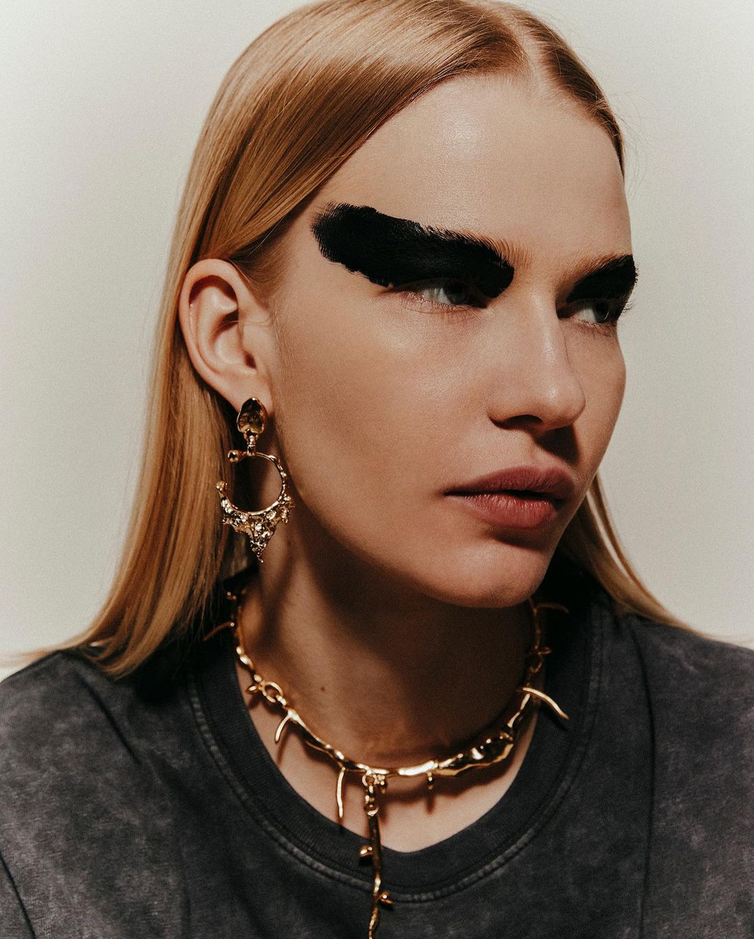 Close-up fashion shot of a blonde woman with bold black eyebrow makeup, wearing gold jewelry against a neutral backdrop.