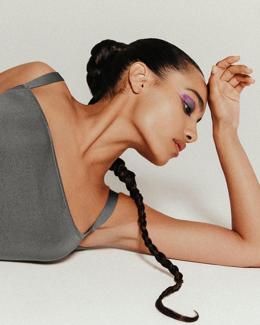 Close-up studio shot of an adult female model with dark braided hair, wearing a grey tank top and bright purple eyeshadow.