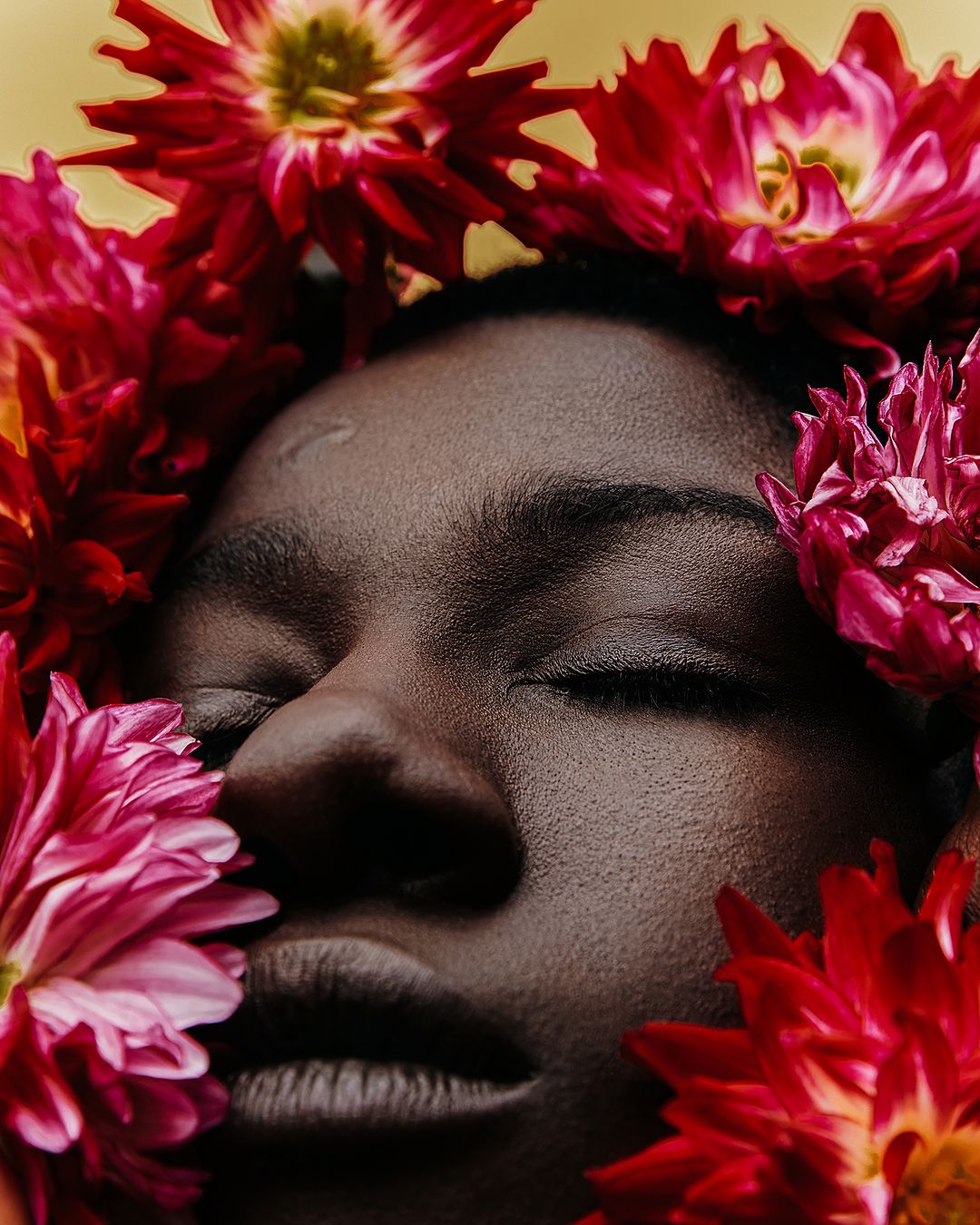 Close-up portrait of an African-American woman with closed eyes surrounded by a vibrant halo of pink and red dahlia flowers.