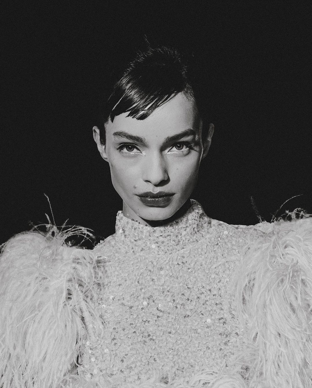 Monochrome close-up of a young adult woman with dark red lipstick wearing a beaded top with feather epaulettes against a dark background.