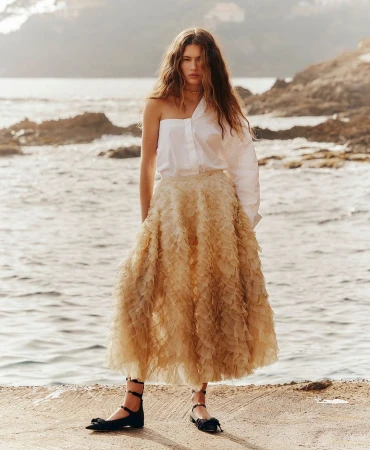 Model standing on a coastal path wearing a white one-shoulder blouse and a beige tiered ruffle skirt against the sea.