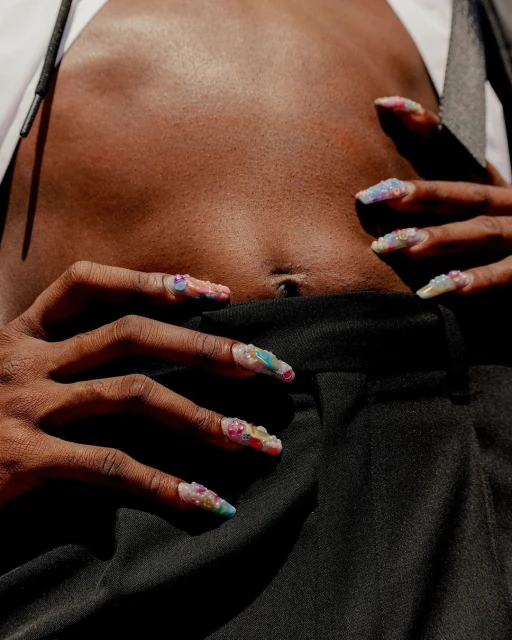 Close-up of a dark-skinned person displaying colorful artificial nails while wearing unbuttoned white shirt and black trousers.