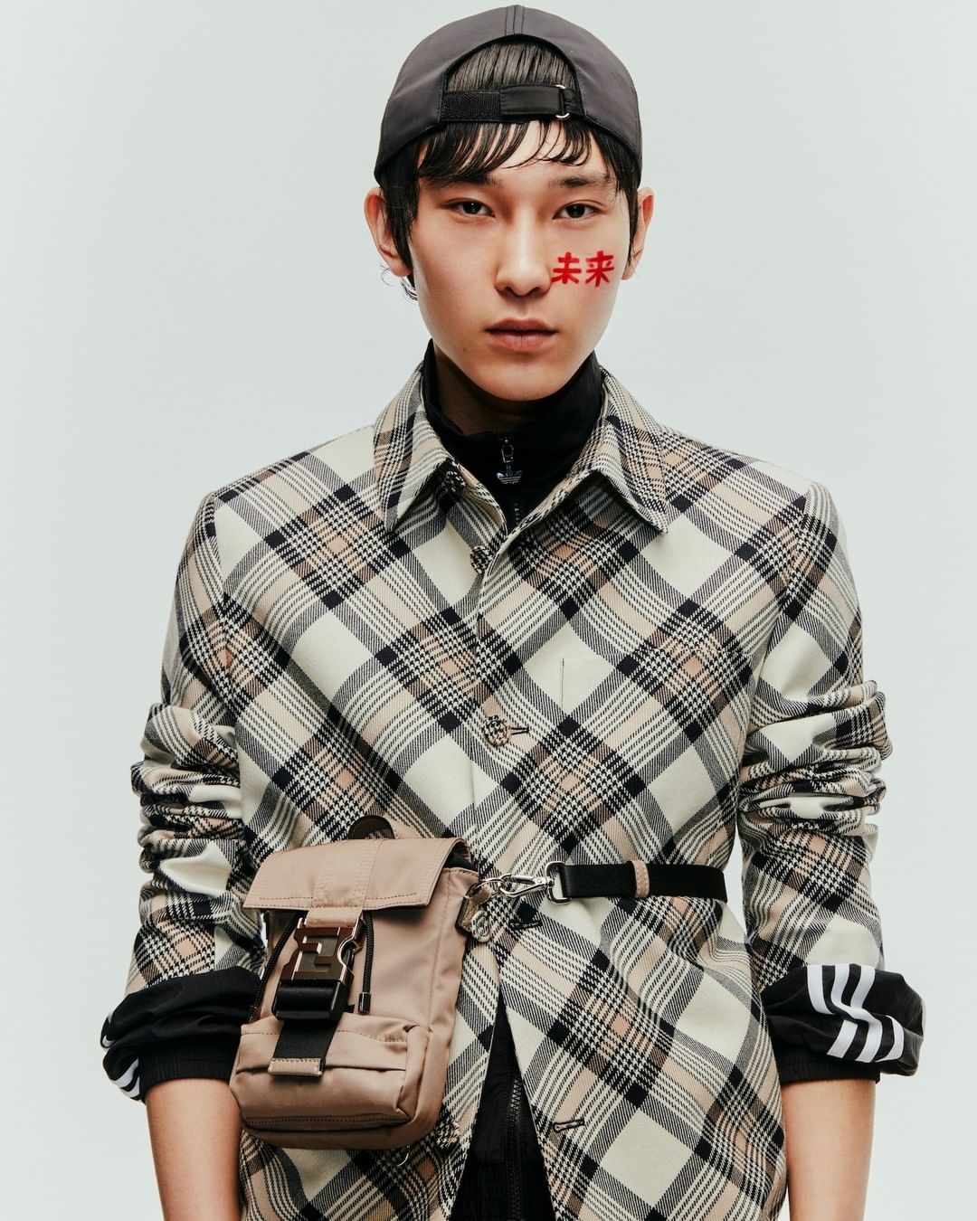 Studio shot of a young adult in a plaid shirt, baseball cap, and tan belt bag, facing forward against a plain background with Chinese characters on their face.