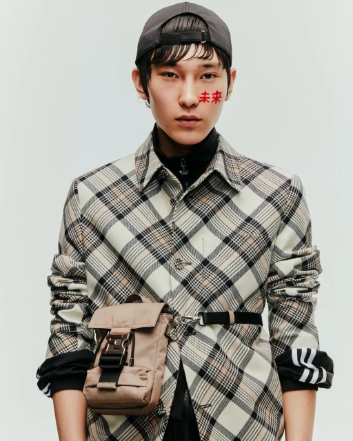 Studio shot of a young adult in a plaid shirt, baseball cap, and tan belt bag, facing forward against a plain background with Chinese characters on their face.
