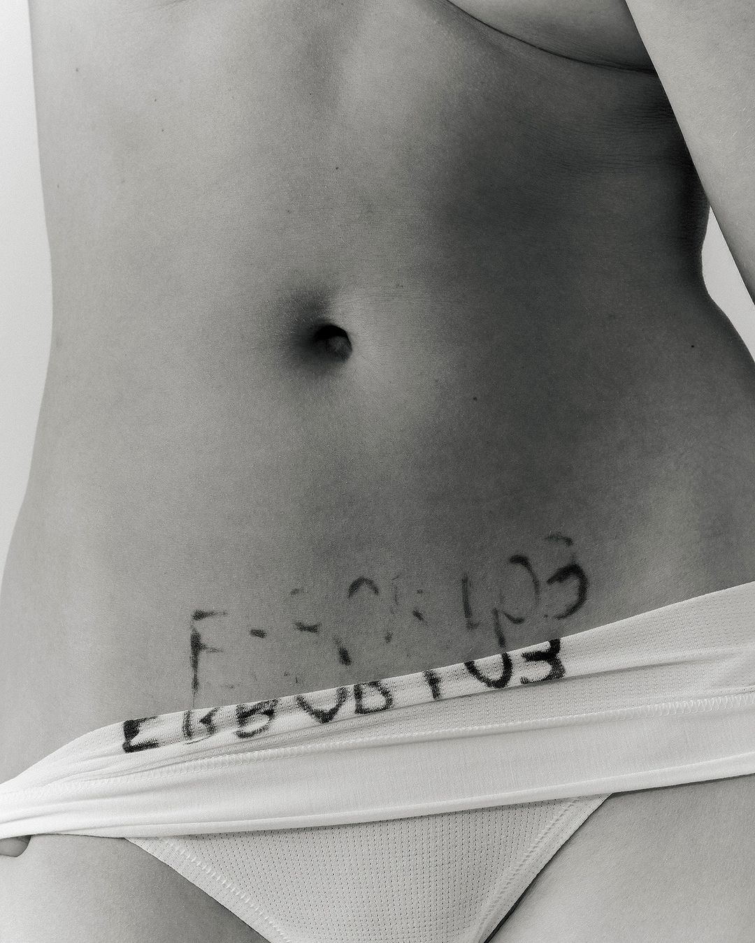 Black and white studio shot of an adult woman's torso, featuring stomach area with written text, and the top of white bikini bottoms.