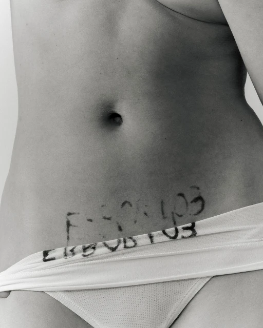 Black and white studio shot of an adult woman's torso, featuring stomach area with written text, and the top of white bikini bottoms.