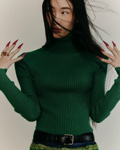 Portrait of a fashionable adult woman with long black hair, wearing a green turtleneck sweater, accessorized with a belt and rings. 