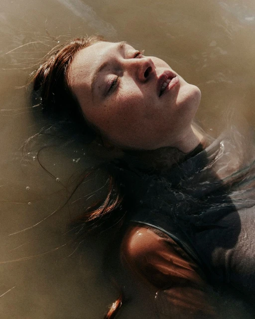 Close-up portrait of a fair-skinned woman with red hair floating in murky water, her eyes closed and face tilted toward the light.