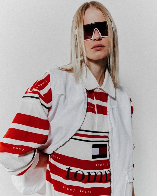 A fair-skinned fashion model poses in a photo studio wearing sunglasses, a striped top with brand logo, and a white vest.