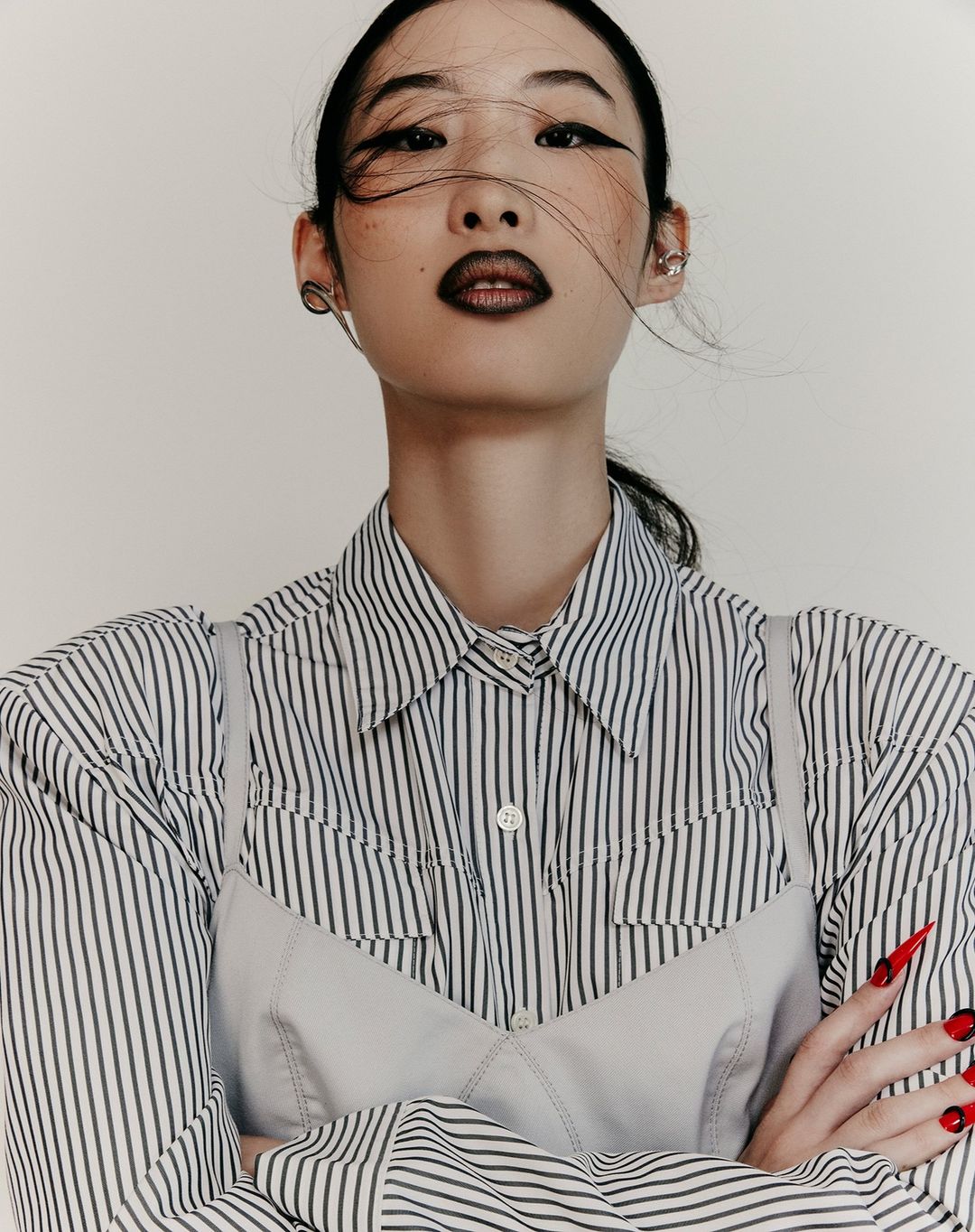 Low angle portrait of an East Asian woman with dark winged eyeliner, dark lips, and a striped shirt under a pale slip top.