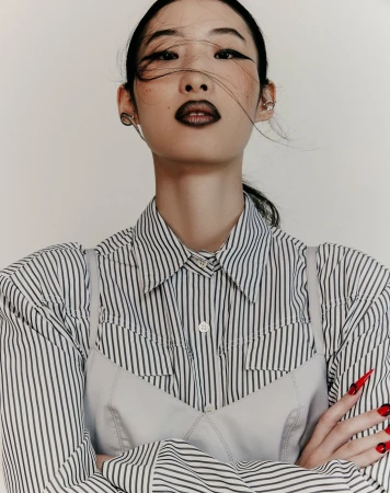 Low angle portrait of an East Asian woman with dark winged eyeliner, dark lips, and a striped shirt under a pale slip top.