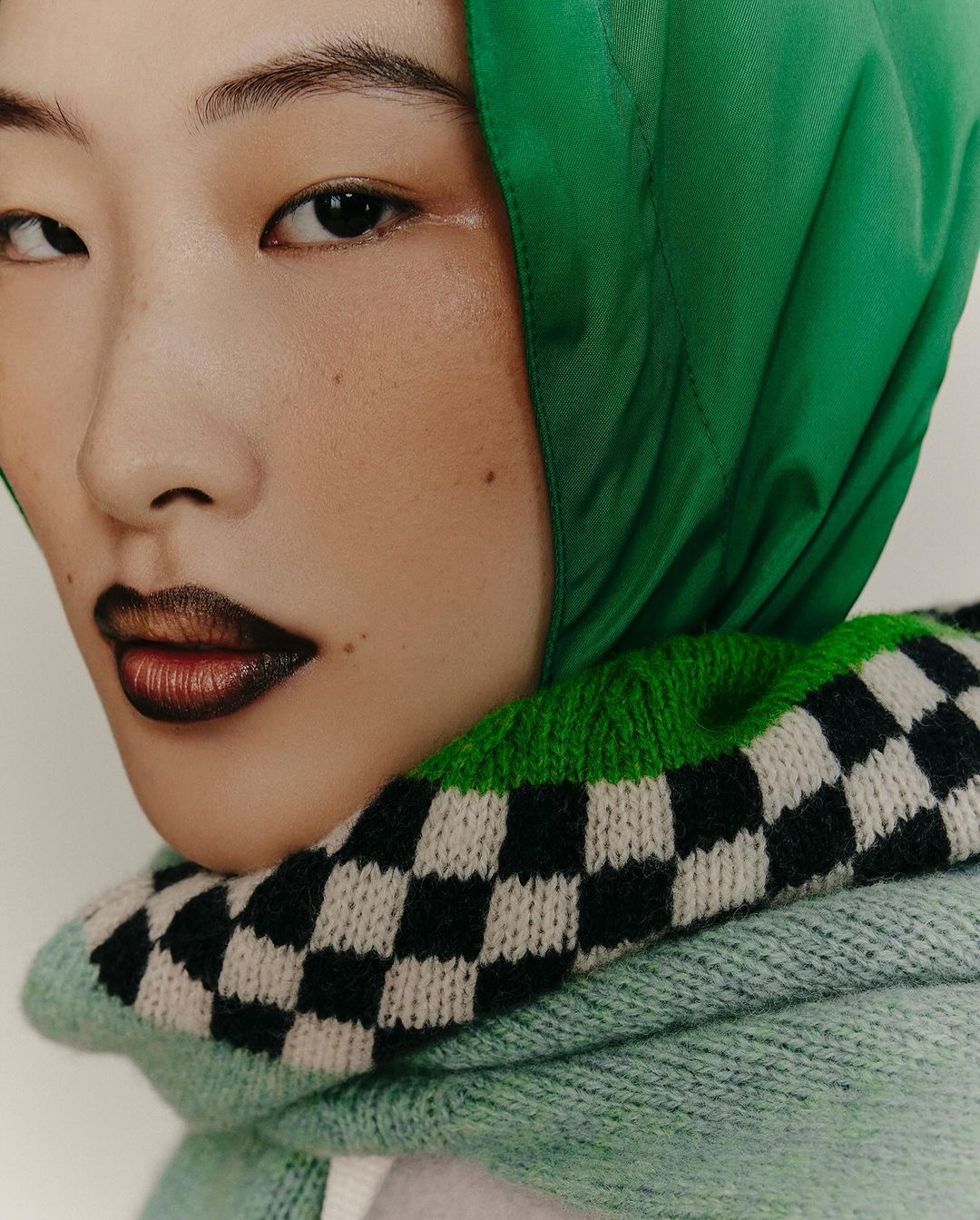 Close-up portrait of an Asian female with dark lipstick wearing a green headscarf and a checkered neck warmer against an off-white background.