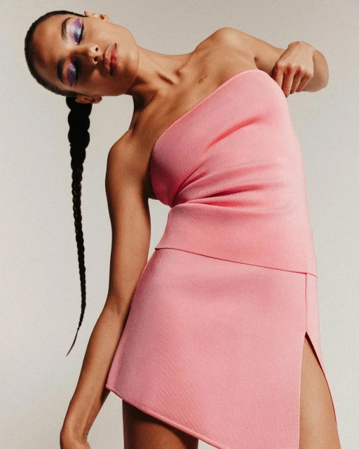 Elegant woman in a pink one-shoulder dress poses in a studio with her head tilted, showcasing contemporary fashion and beauty in simple setting.