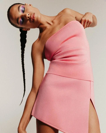 Elegant woman in a pink one-shoulder dress poses in a studio with her head tilted, showcasing contemporary fashion and beauty in simple setting.