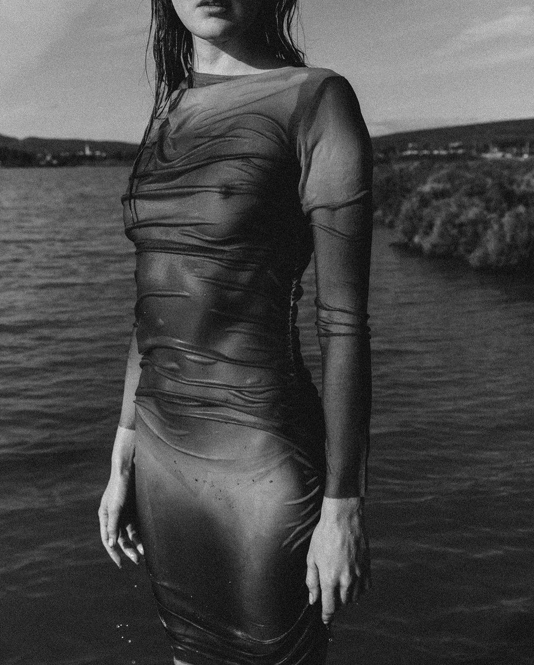 Monochrome portrait of a woman in a wet sheer dress standing in water, featuring soft light and a distant shoreline.