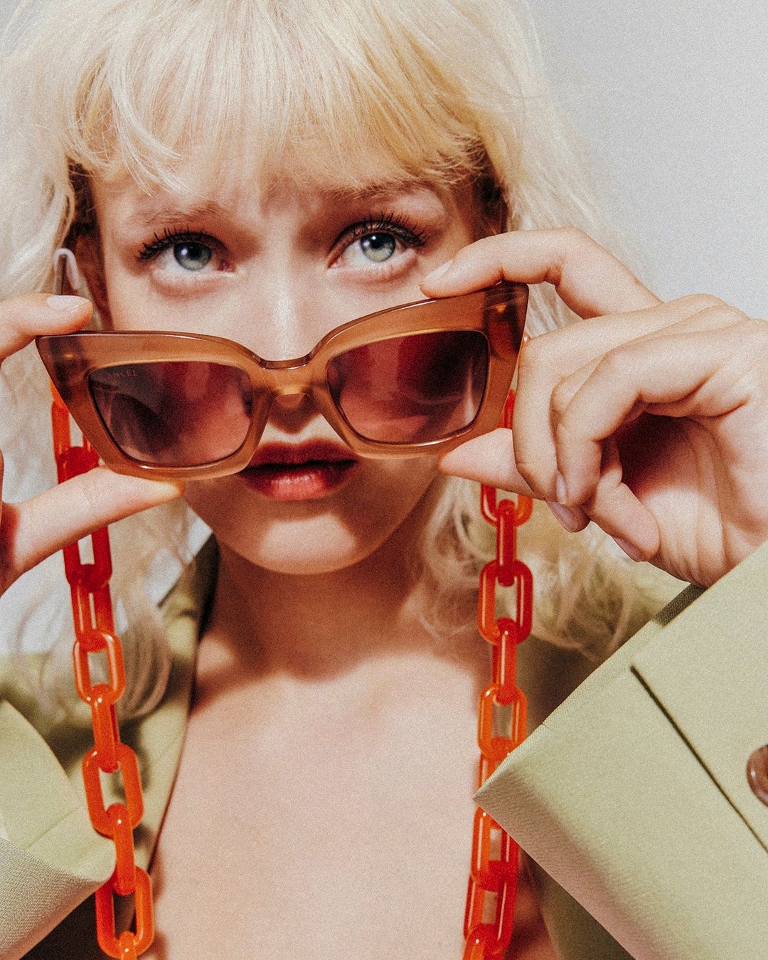 Close-up shot of a blonde woman holding brown sunglasses with an orange chain, showcasing a modern and fashionable aesthetic.