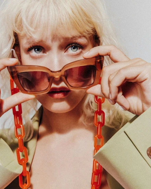 Close-up shot of a blonde woman holding brown sunglasses with an orange chain, showcasing a modern and fashionable aesthetic.